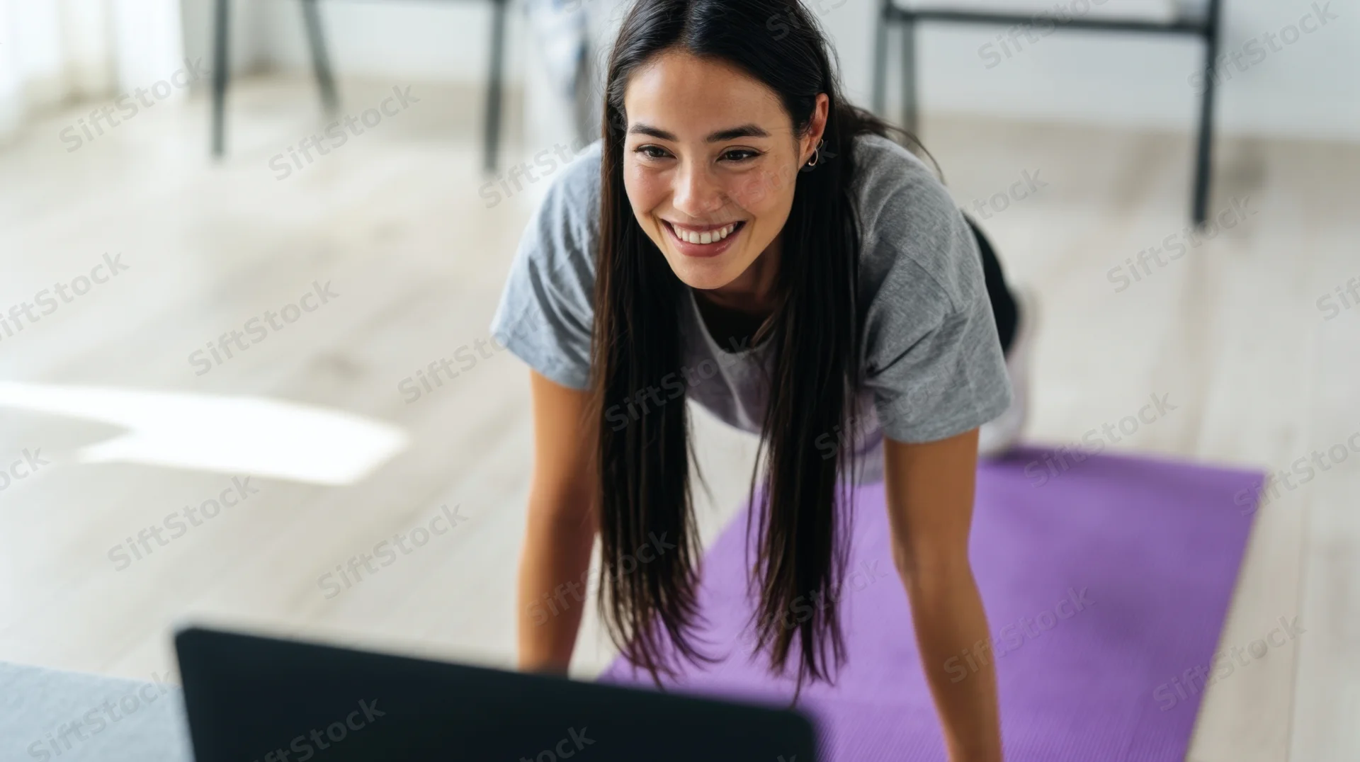 Woman doing plank exercise on yoga mat while smiling at laptop screen at home