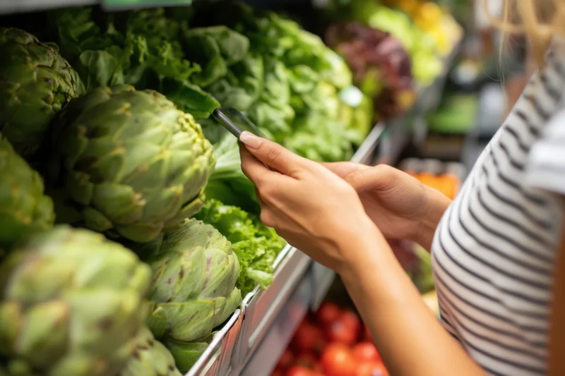 Woman using smartphone while selecting vegetables in grocery store produce section