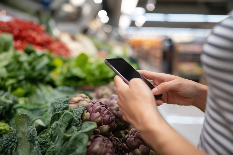 Woman using smartphone while shopping for fresh vegetables in a supermarket produce aisle