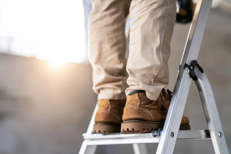 Worker standing on aluminum ladder wearing work boots and trousers outdoors in sunlight