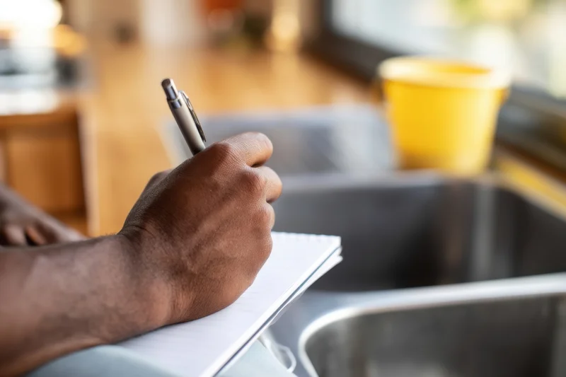 Man writing in notebook at kitchen sink with pen in hand