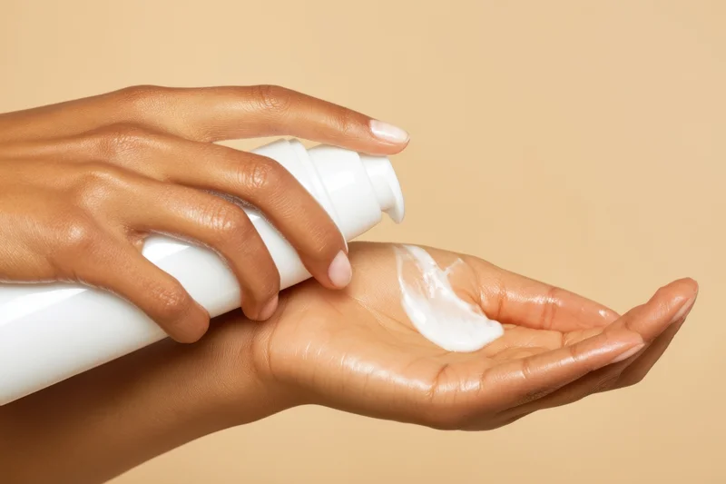 Woman dispensing white cream from a pump bottle into her palm against a beige background