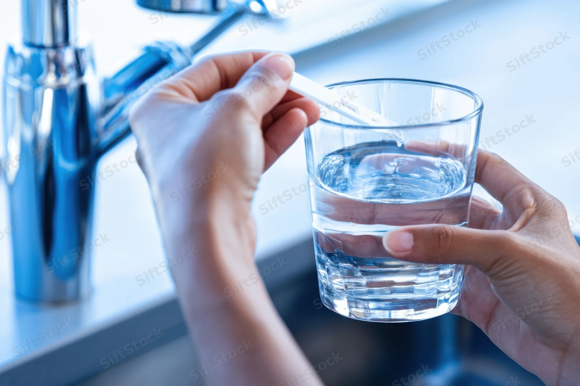 Hands holding a glass of water and inserting a test strip near a kitchen faucet
