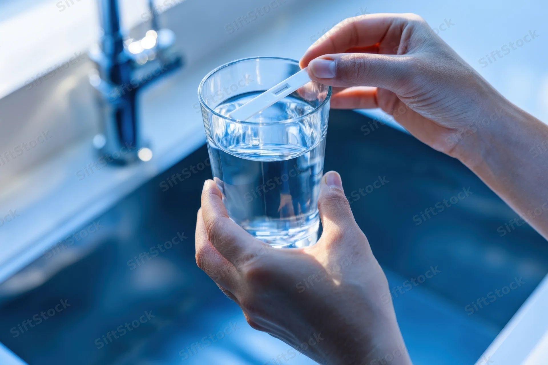 Hands holding a glass of water with a test strip inserted over a kitchen sink