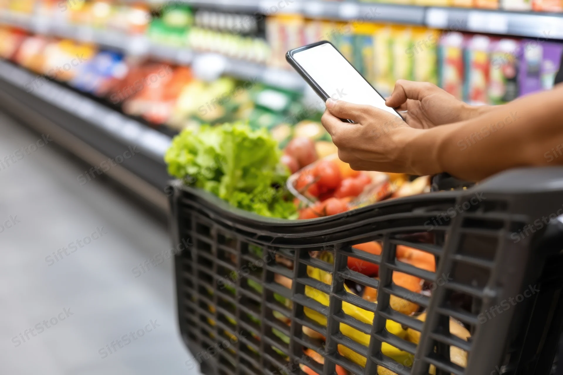Person holding smartphone over grocery basket filled with fresh produce in supermarket aisle