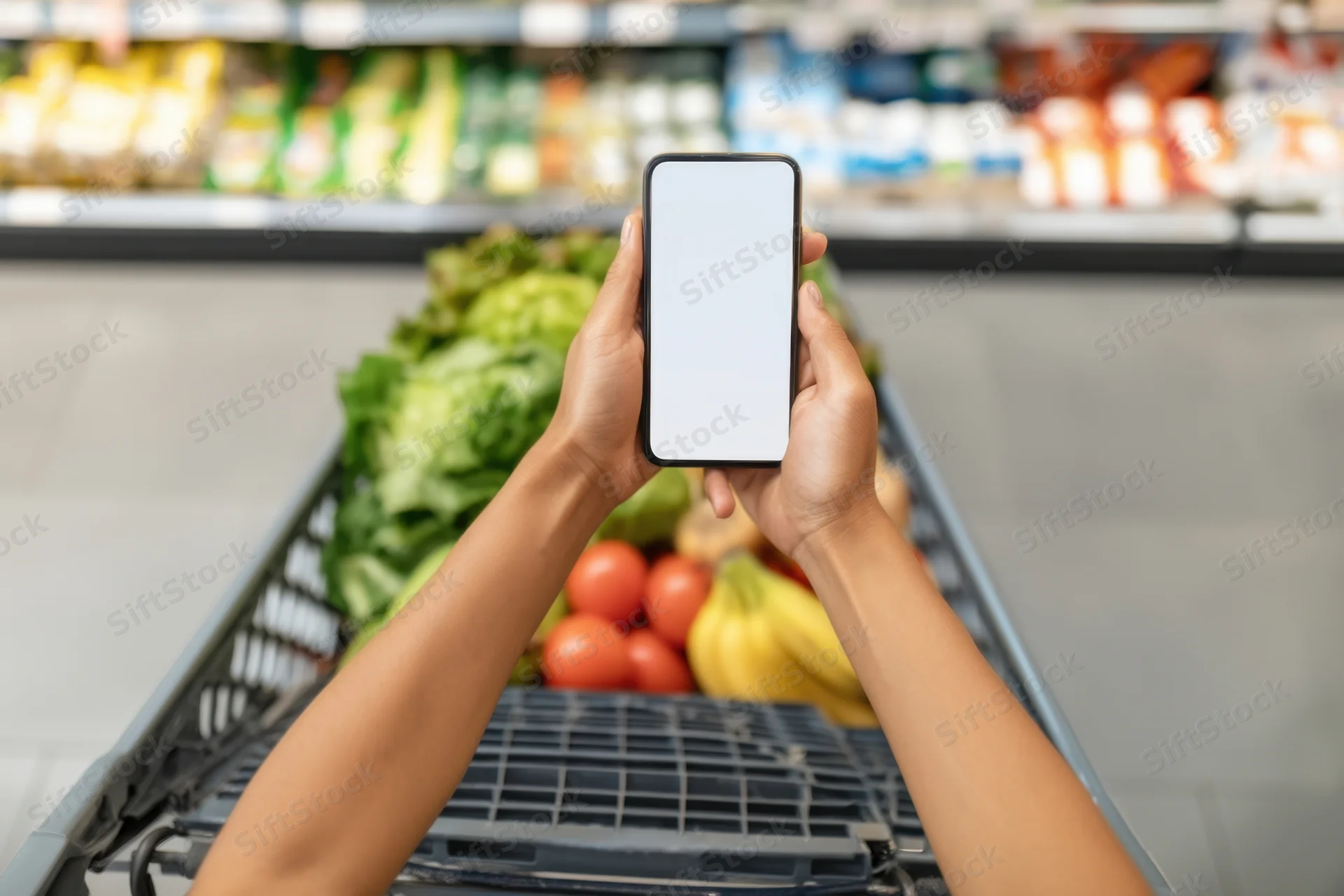 Woman holding smartphone over shopping cart filled with fresh produce in supermarket