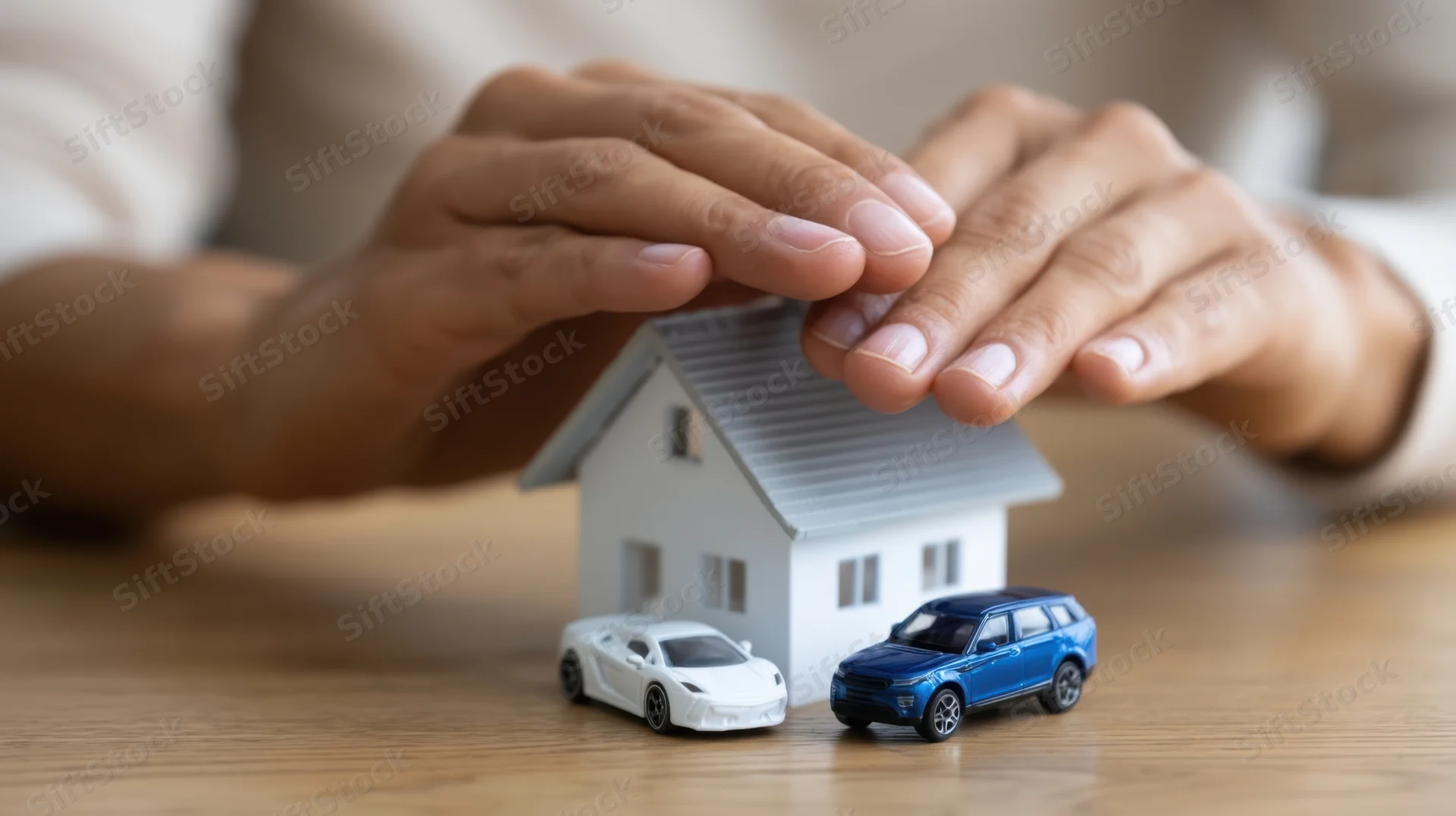 Hands covering a miniature house and toy cars on a wooden table
