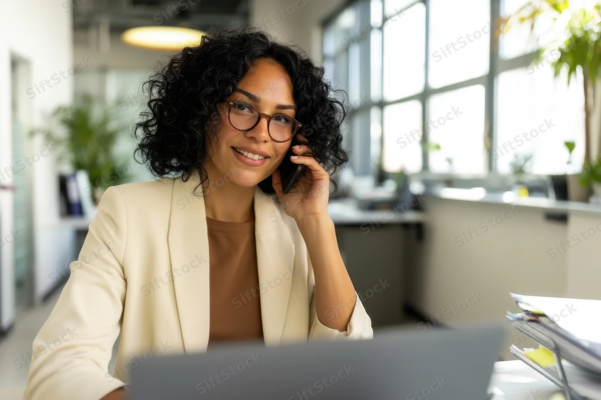 Woman in beige blazer smiling while on phone call at office desk with laptop and window view