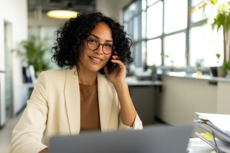 Woman in beige blazer smiling while on phone call at office desk with laptop and window view