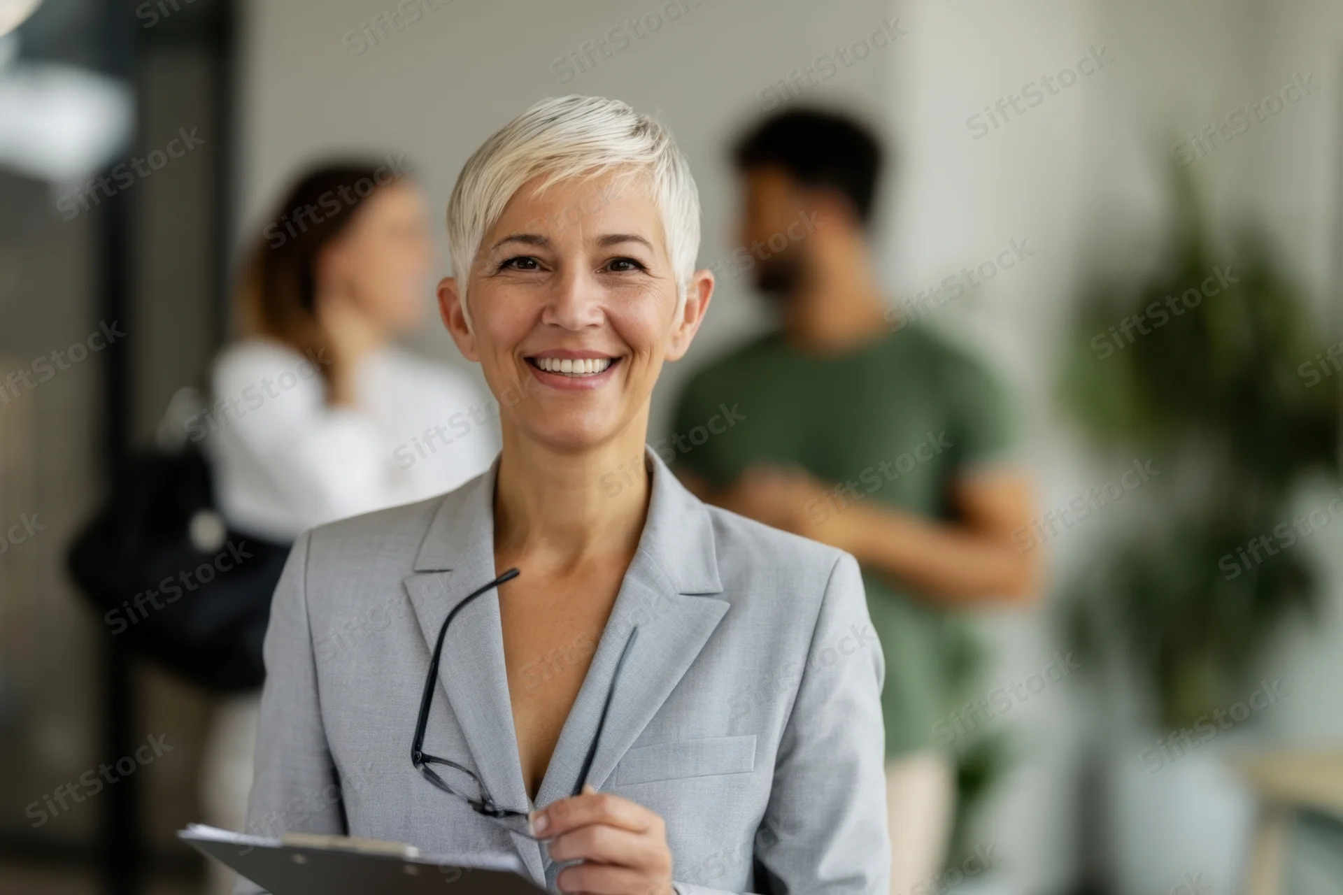Smiling woman in gray suit holding clipboard in office with colleagues in background