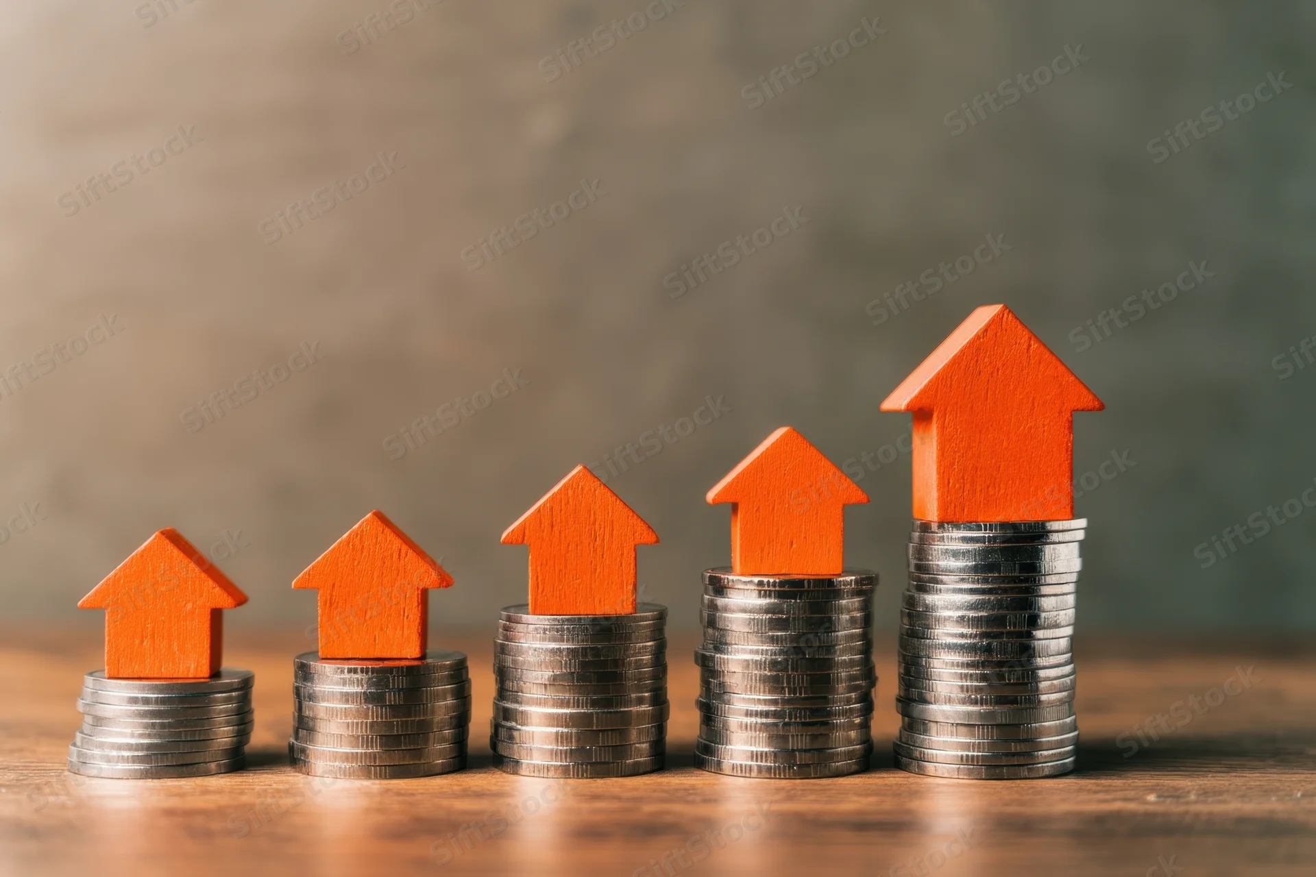 Five orange wooden houses stacked on rising piles of silver coins on a wooden surface
