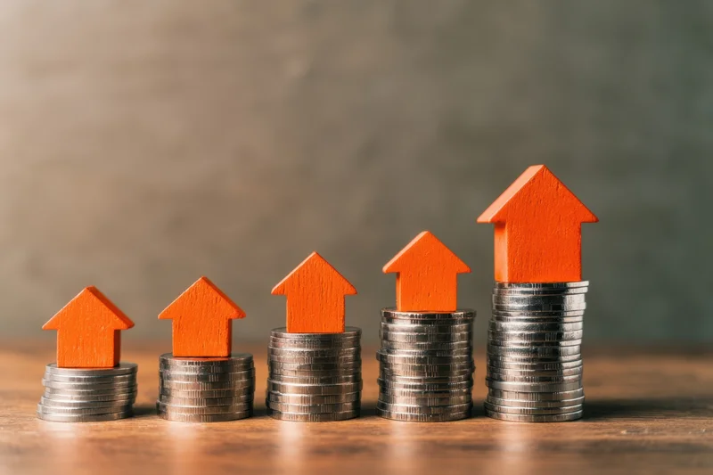 Five orange wooden houses stacked on rising piles of silver coins on a wooden surface