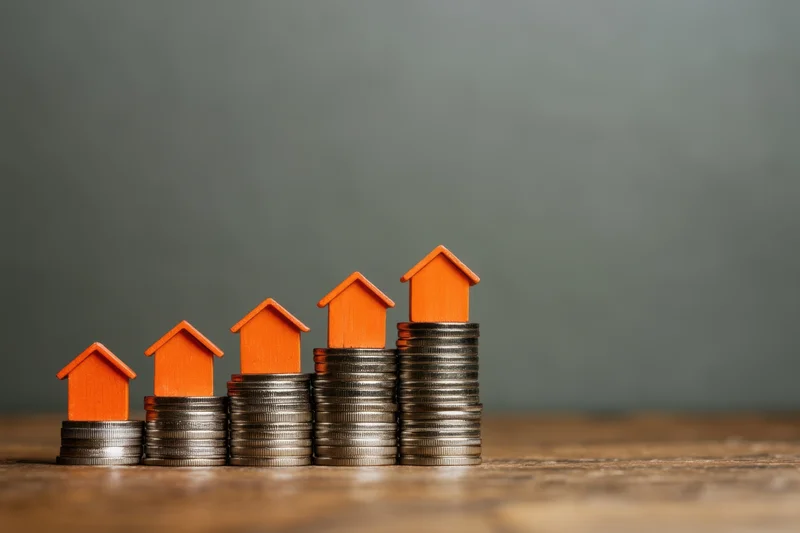 Five orange toy houses stacked on increasing piles of silver coins on a wooden surface