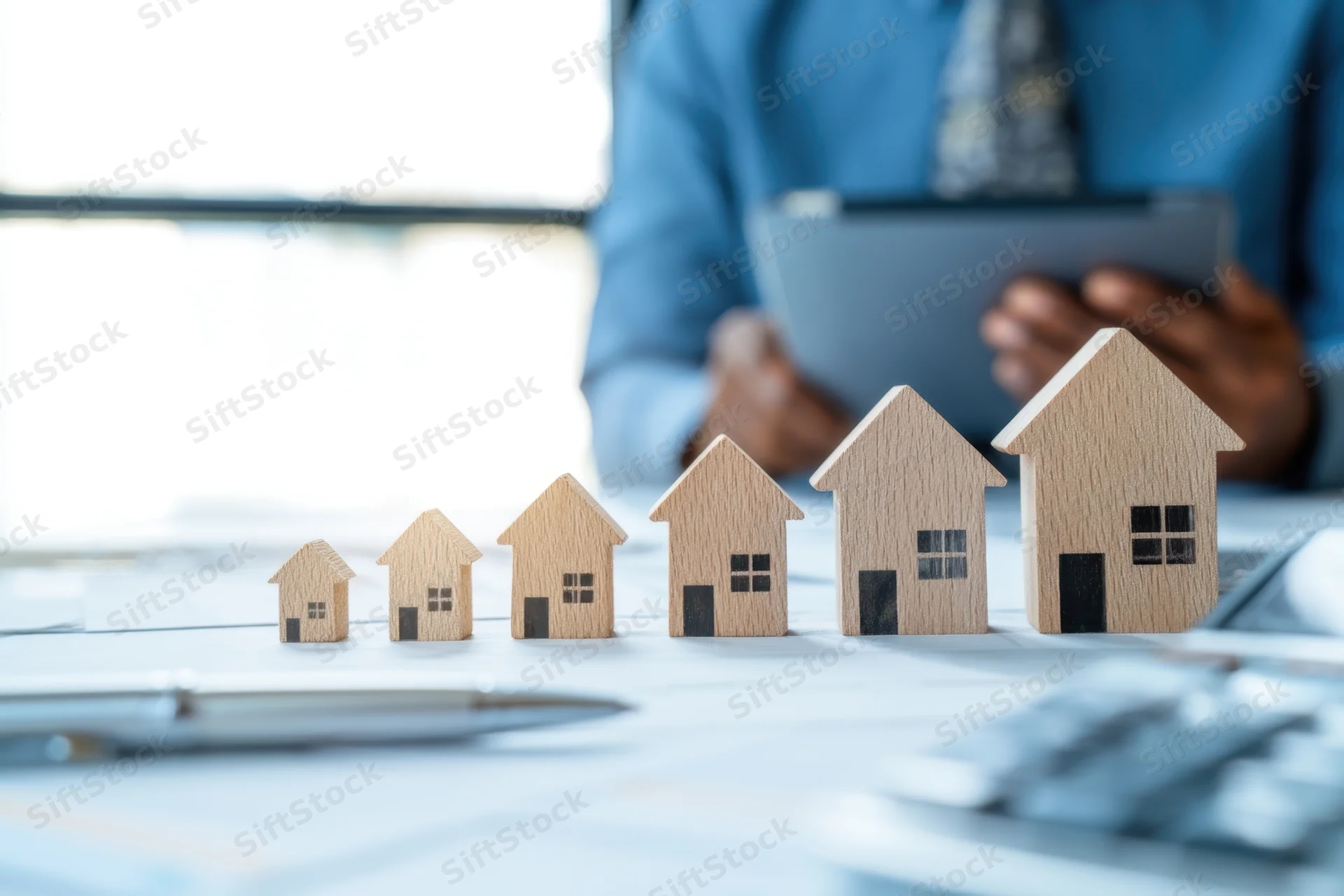 Wooden house models arranged in increasing size on a desk with a person using a tablet in the background