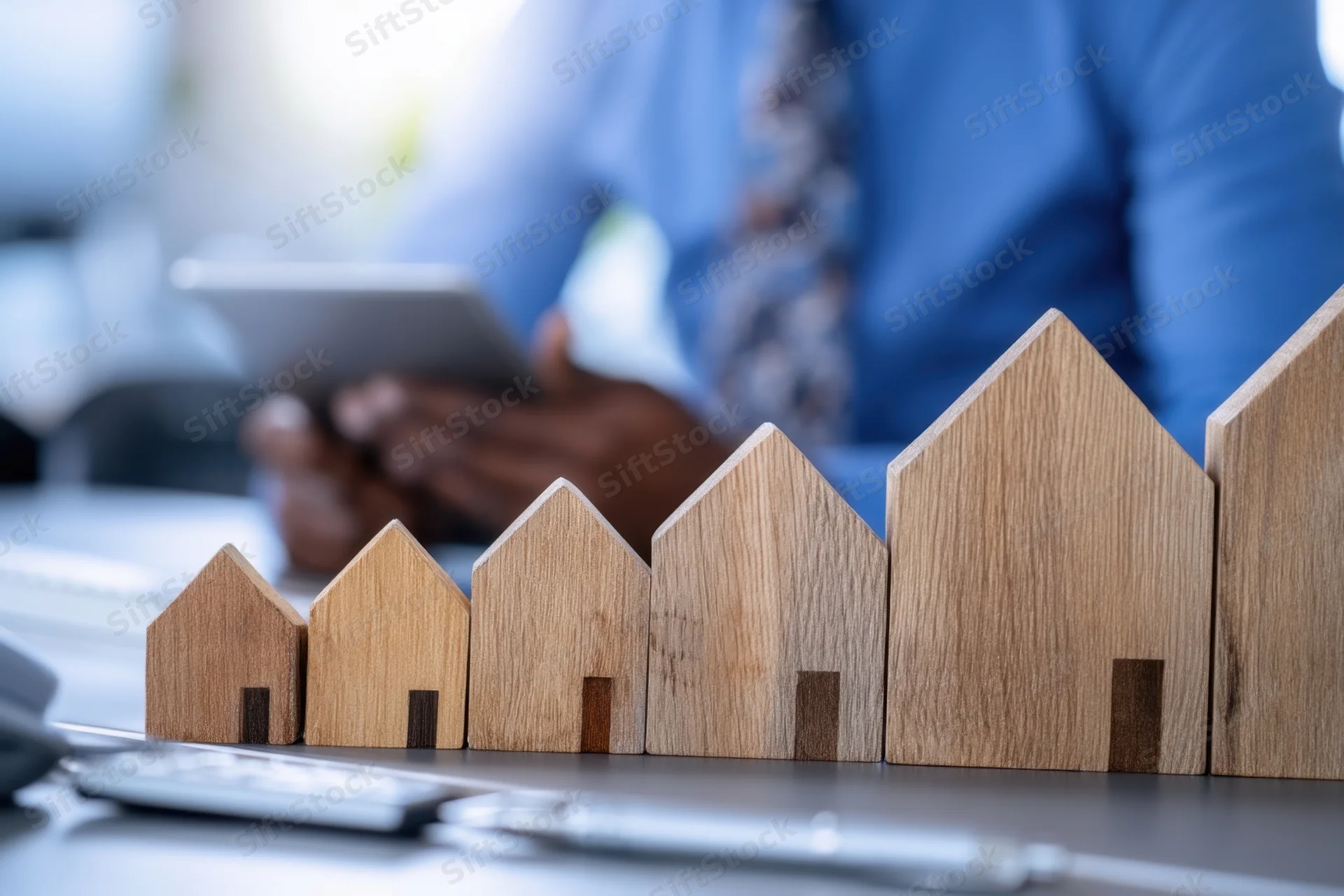 Wooden house models arranged in increasing size on a desk with a man using a tablet in the background