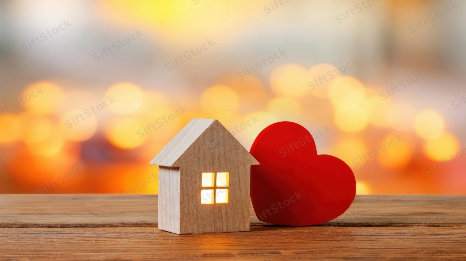 Small wooden house with glowing window and red heart on wooden surface against warm bokeh background