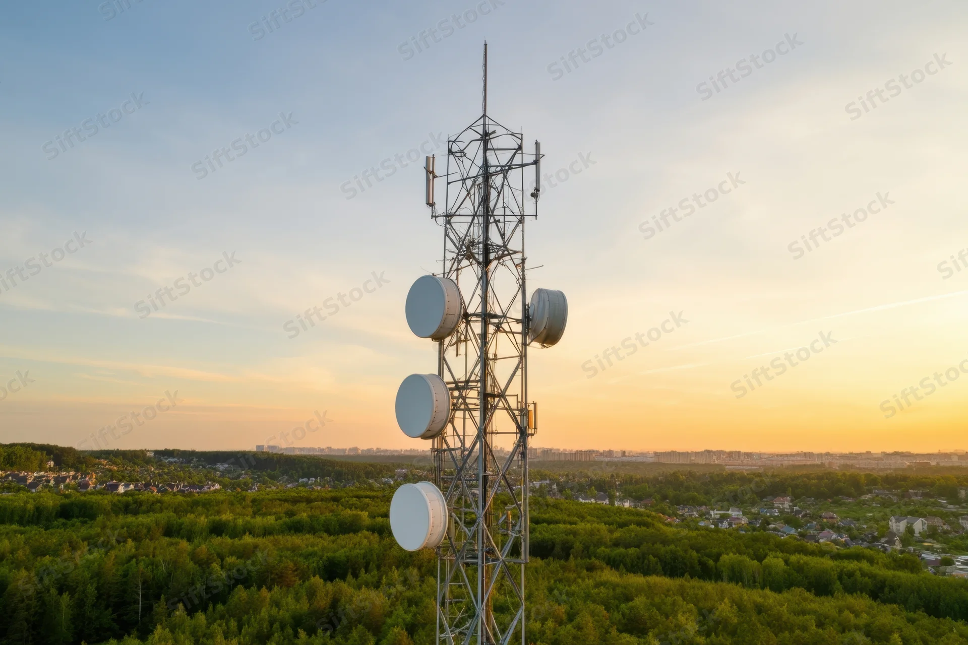 Telecommunications tower with multiple antennas and satellite dishes rising above a forested landscape at sunset, overlooking a city in the distance