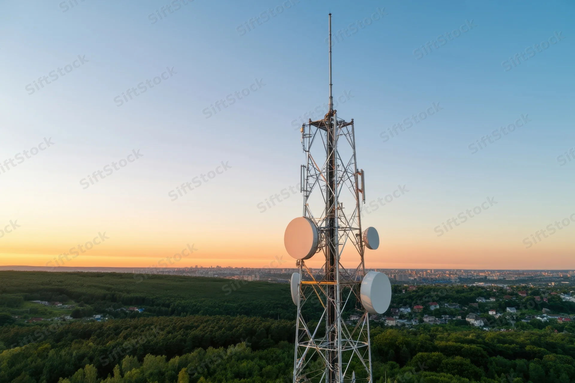 Telecommunications tower with multiple antennas and dishes overlooking a city and forest at sunset