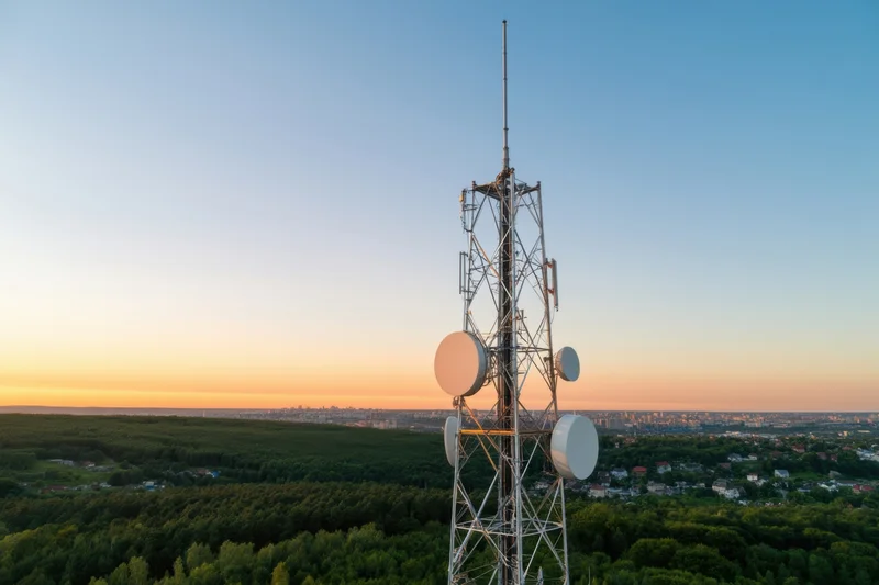 Telecommunications tower with multiple antennas and dishes overlooking a city and forest at sunset