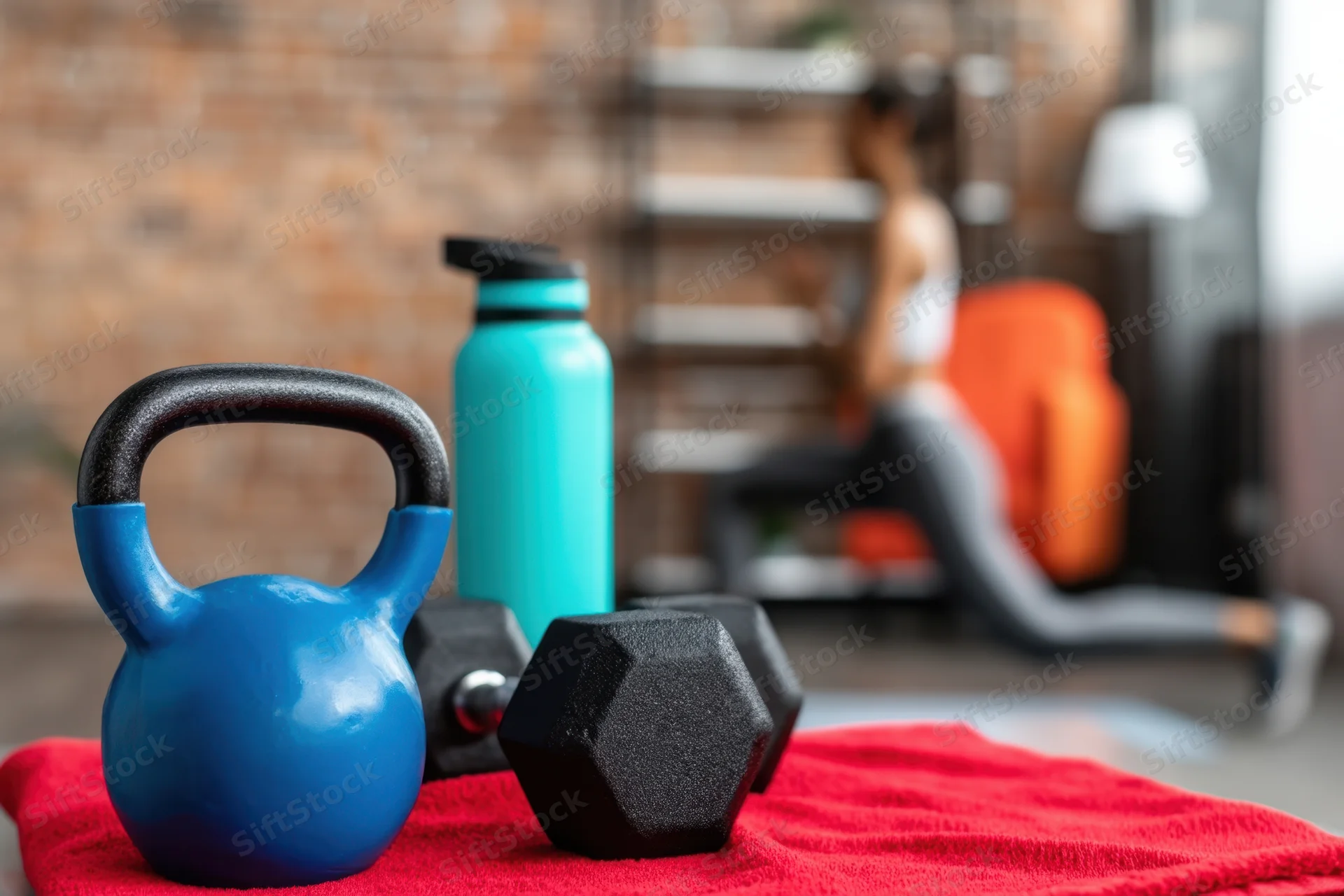 Blue kettlebell and black dumbbells on red towel with water bottle, woman exercising in background at home gym