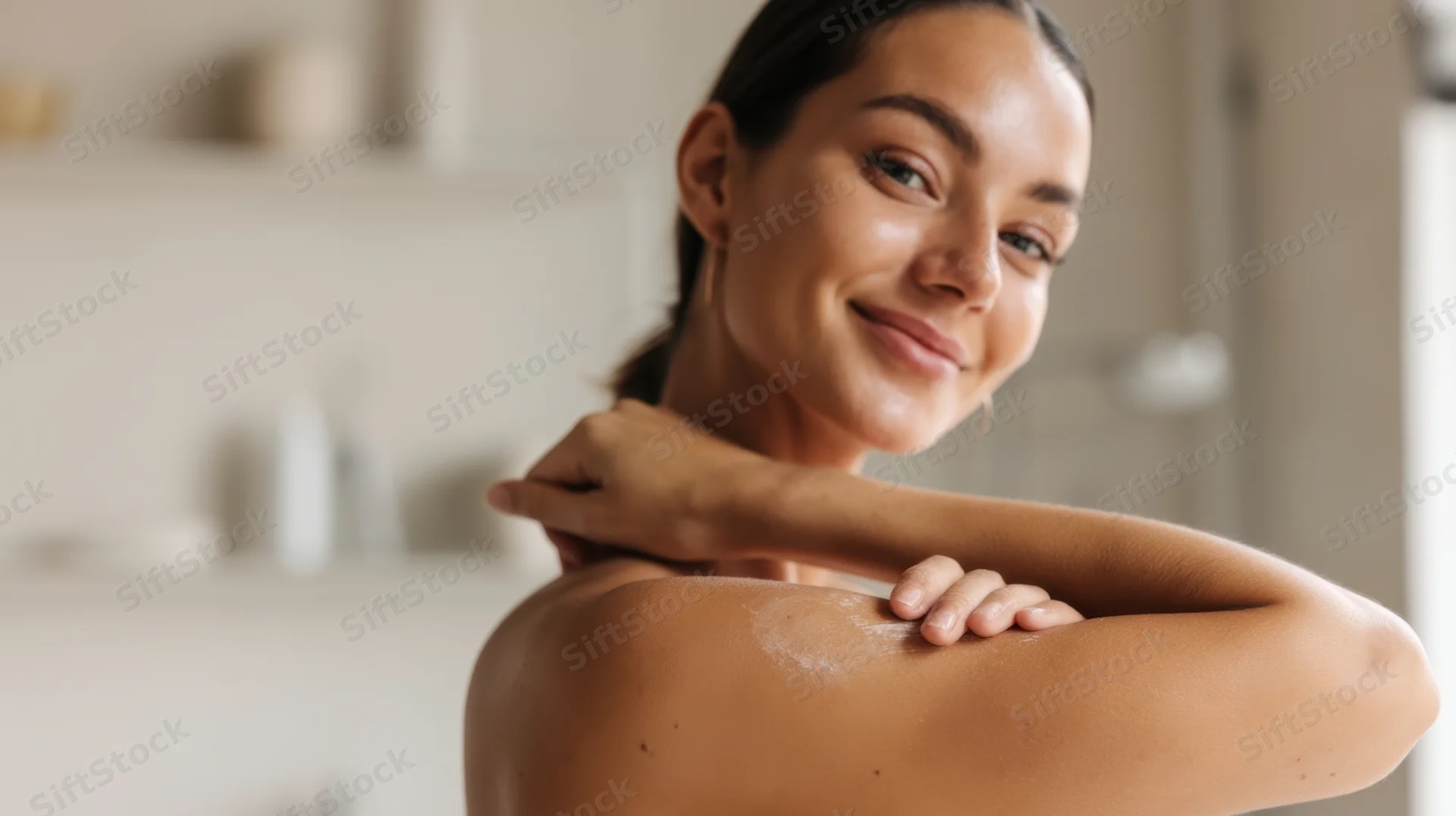 Woman smiling while applying moisturizer to her shoulder in a bathroom