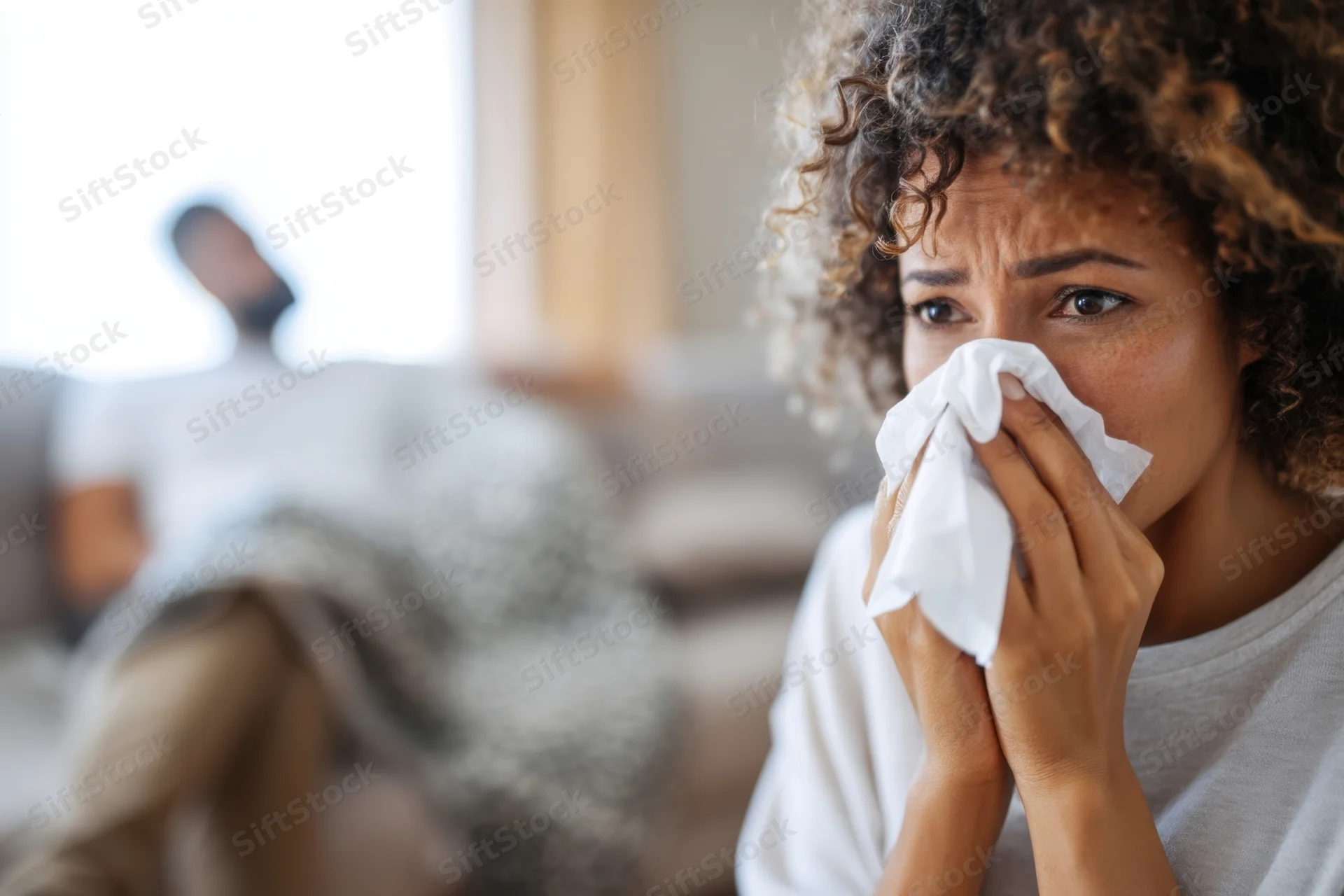 Woman holding tissue to her nose while showing signs of illness in a living room with a man resting on a sofa in the background