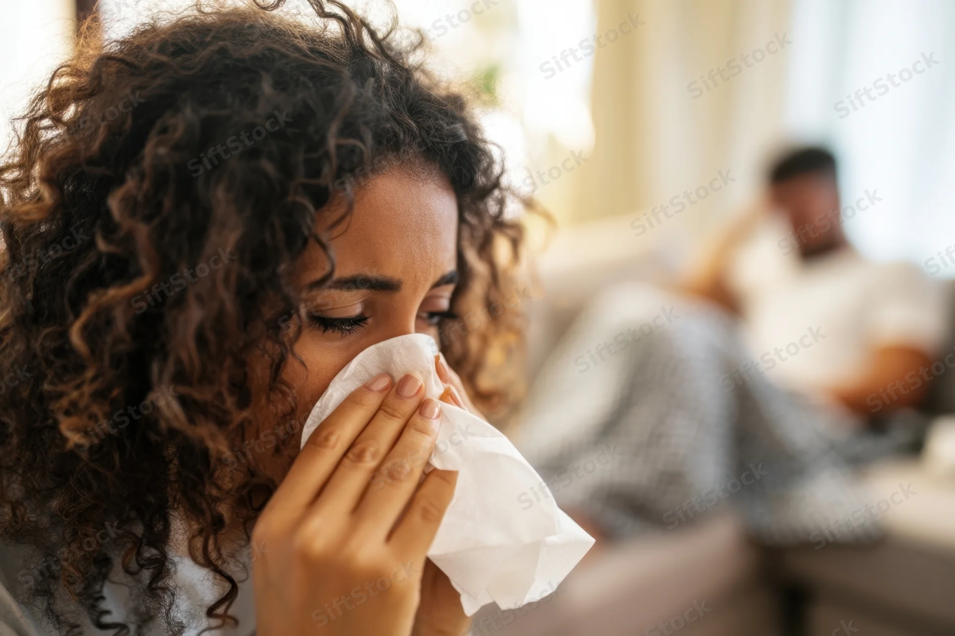 Woman blowing her nose with a tissue while resting at home, with a man in the background on the couch