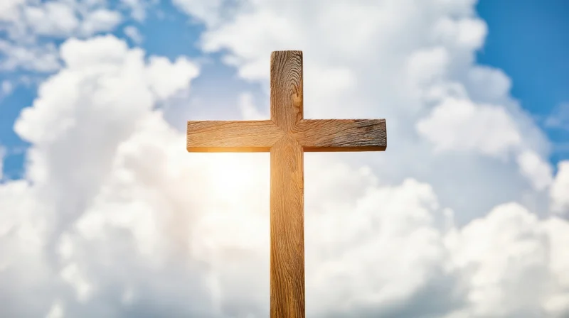 Wooden cross standing against a bright blue sky with white clouds and sunlight behind it