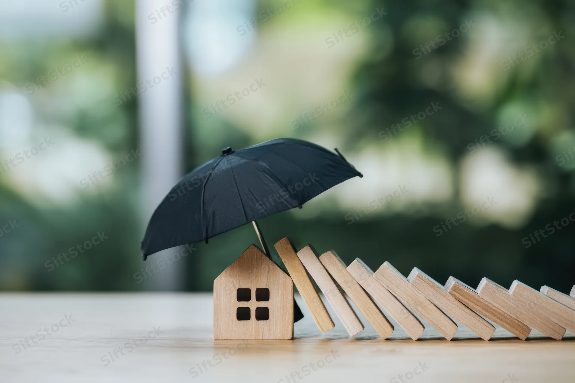 Wooden house model under black umbrella shielding it from falling dominoes on a table