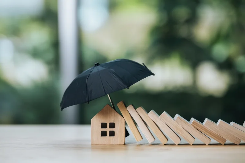 Wooden house model under black umbrella shielding it from falling dominoes on a table