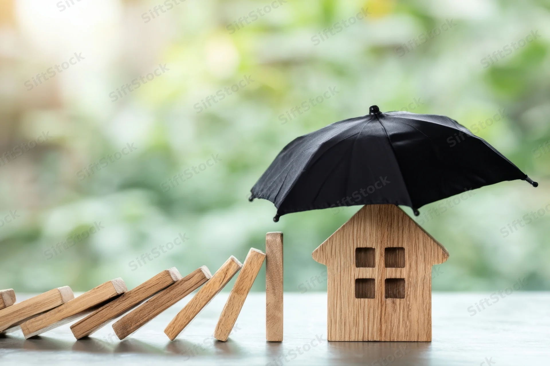 Wooden house model under black umbrella with falling dominoes on table