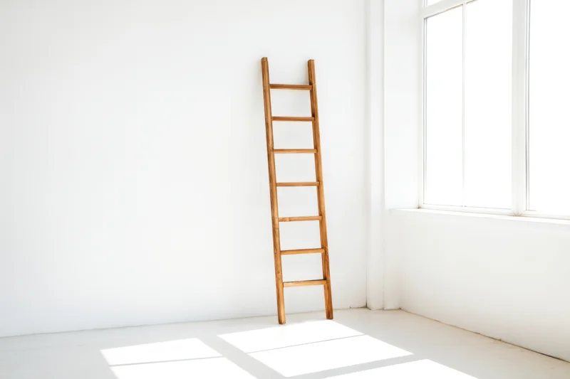 Wooden ladder leaning in a white minimalist room corner near a window with sunlight casting shadows on the floor