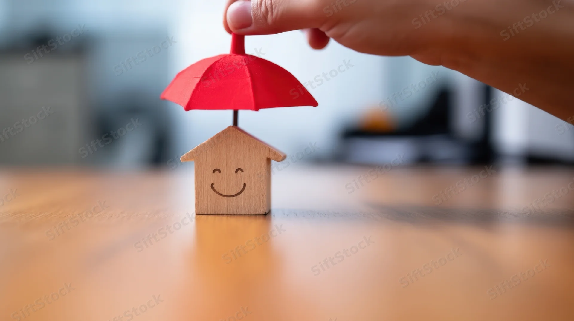 Hand holding red umbrella over smiling wooden house model on desk
