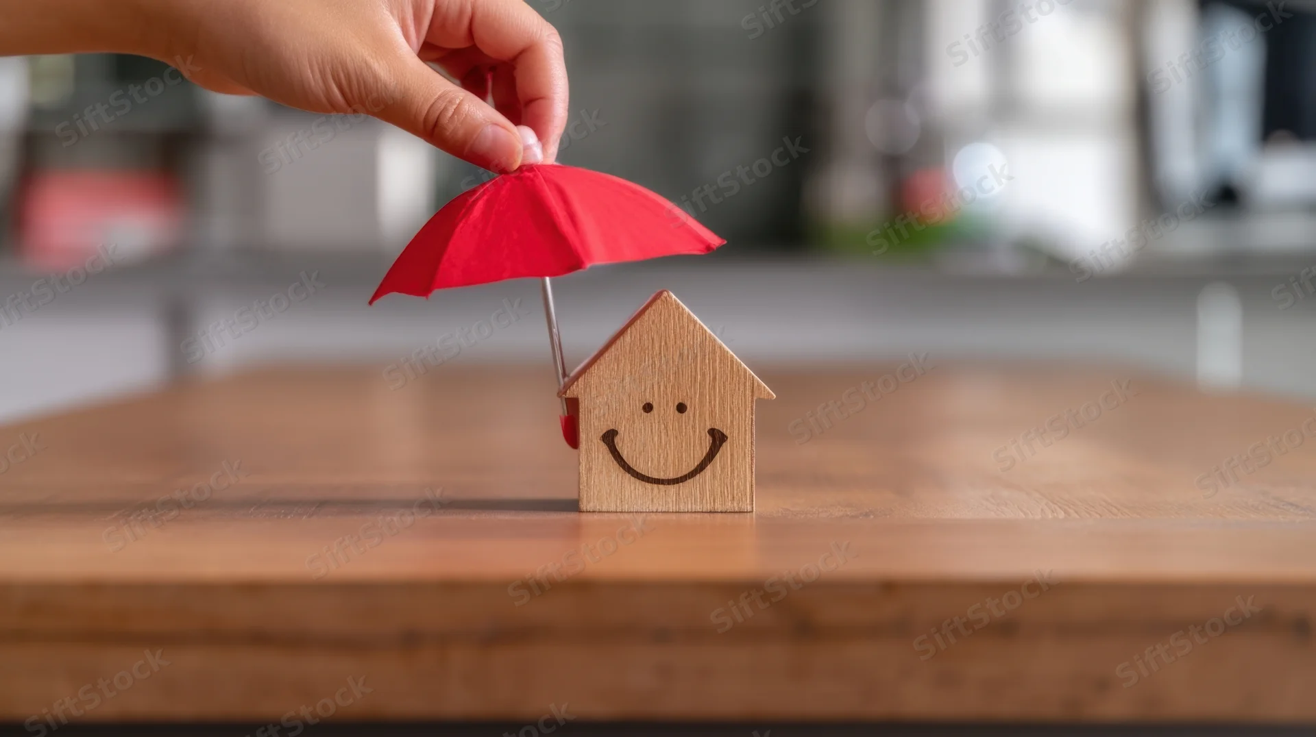 Hand holding red umbrella over smiling wooden house model on wooden table