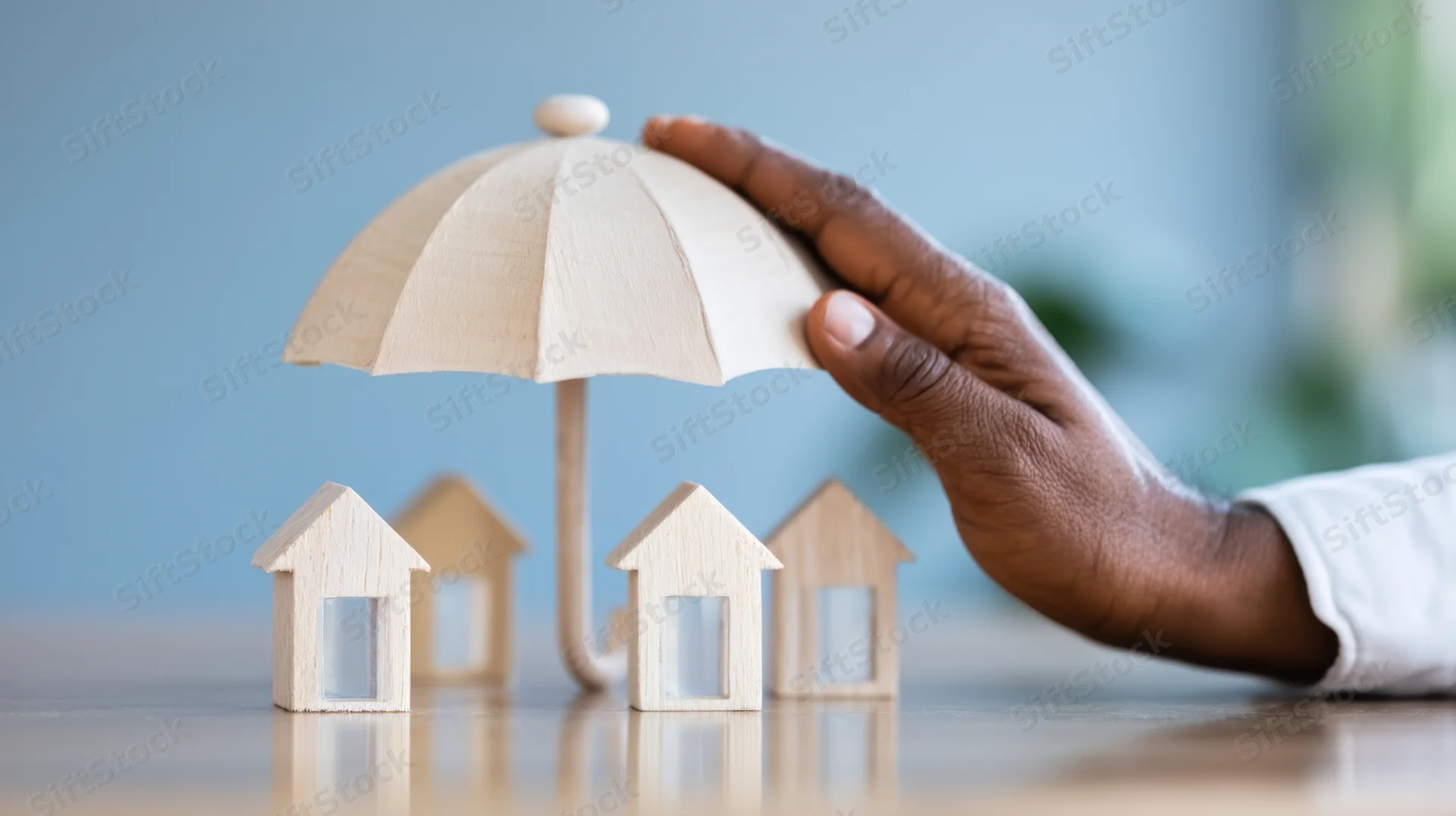 Hand holding wooden umbrella over miniature houses on desk