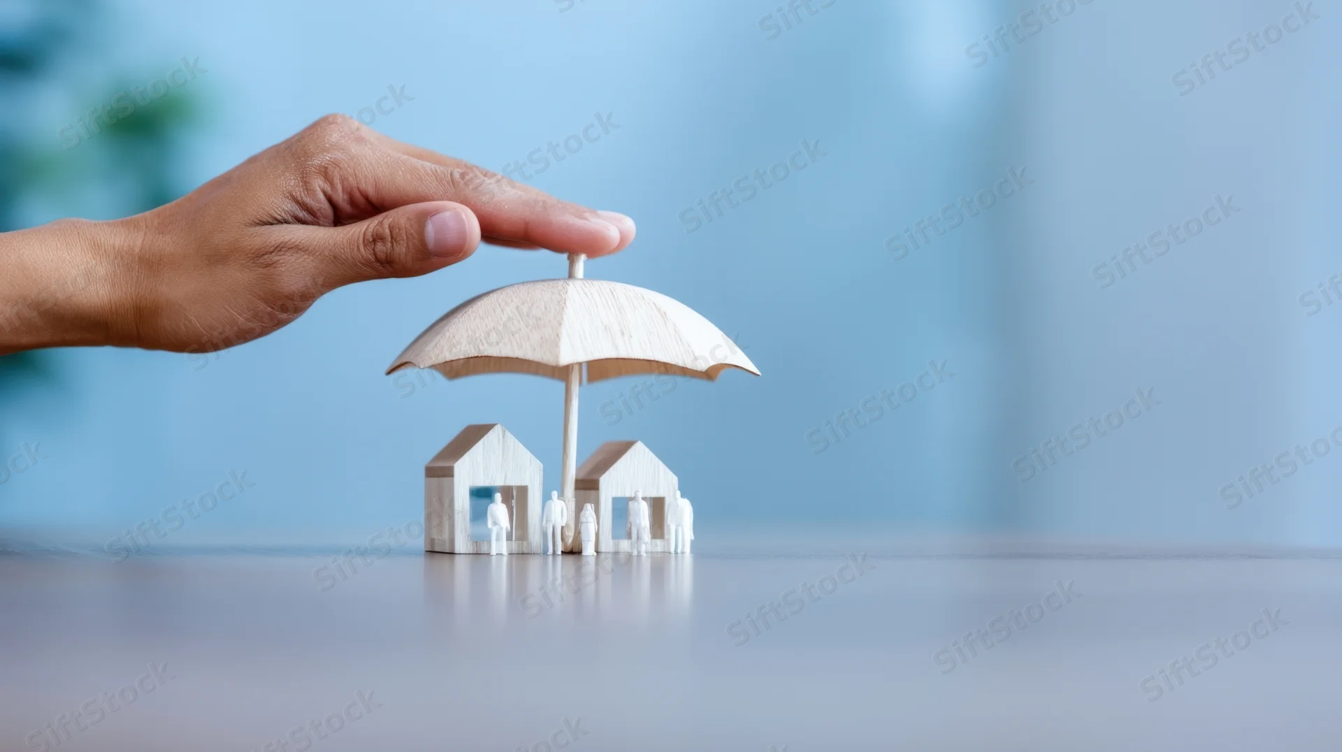 Hand holding wooden umbrella over miniature houses and figures on a desk