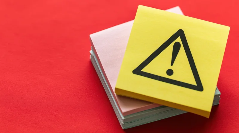 Yellow sticky note with warning triangle symbol on red background, stacked with other notes