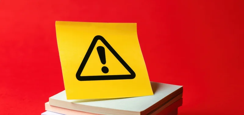 Yellow warning symbol on sticky note placed on stack of books against red background