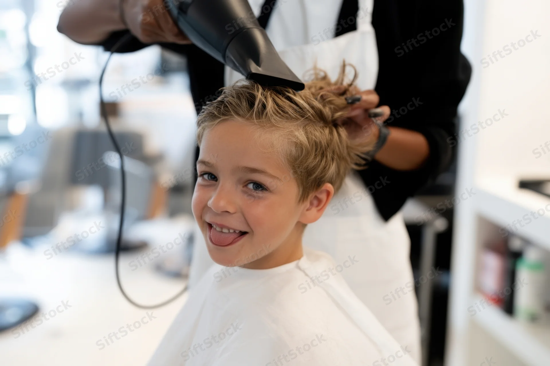 Young boy smiling with tongue out while stylist dries his hair in salon