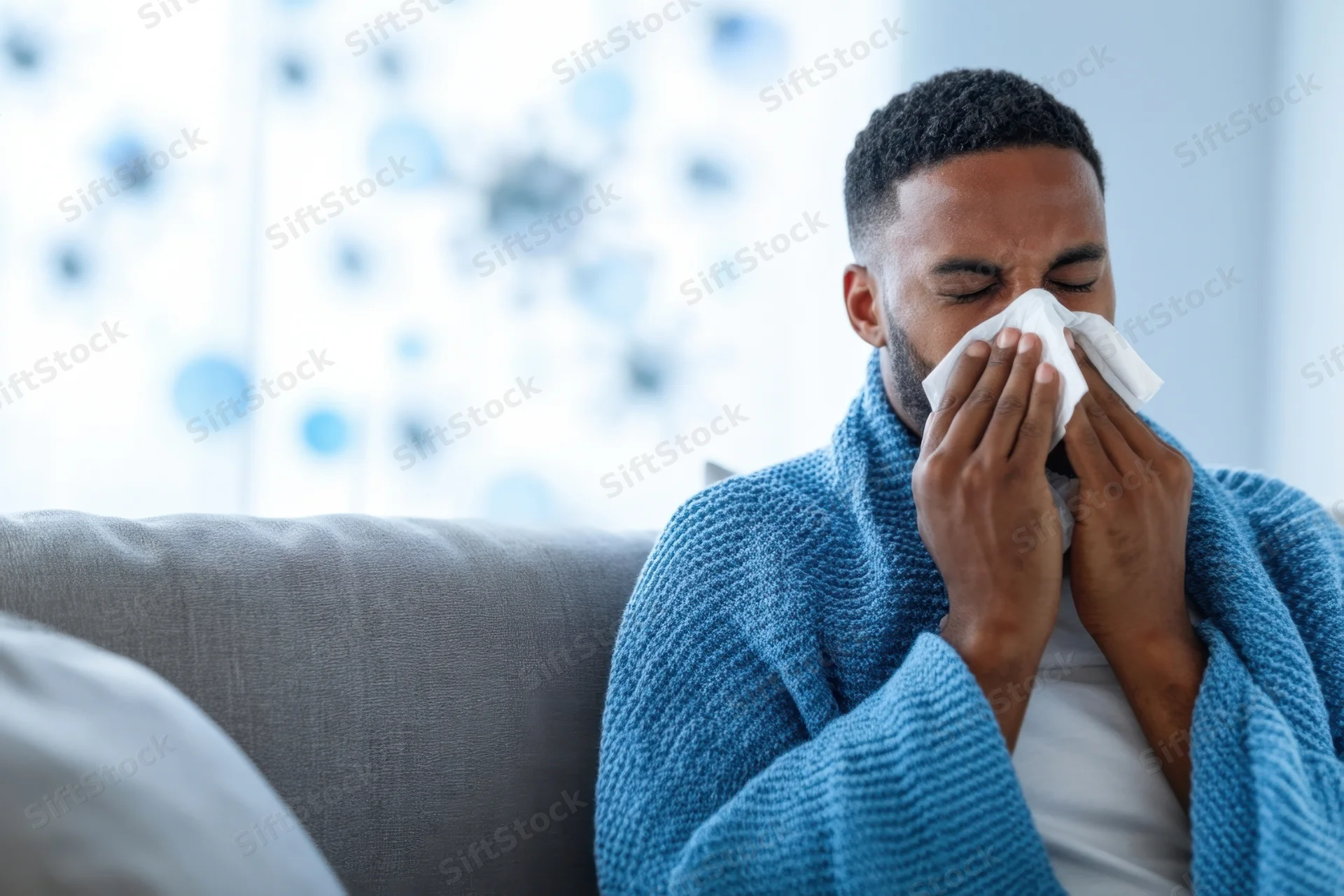 Young man sneezing into tissue while wrapped in blue robe on sofa