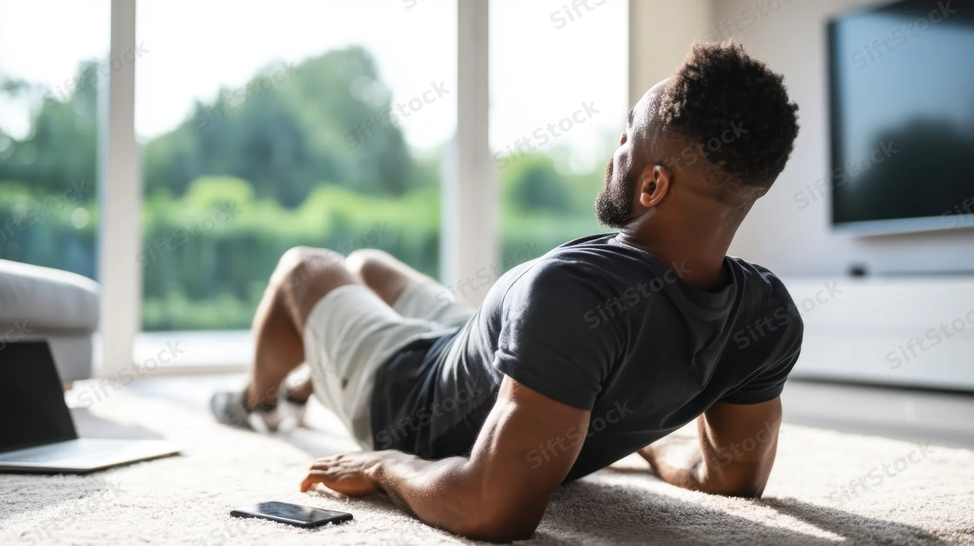 Young man doing pushups on carpet in living room with laptop and smartphone nearby
