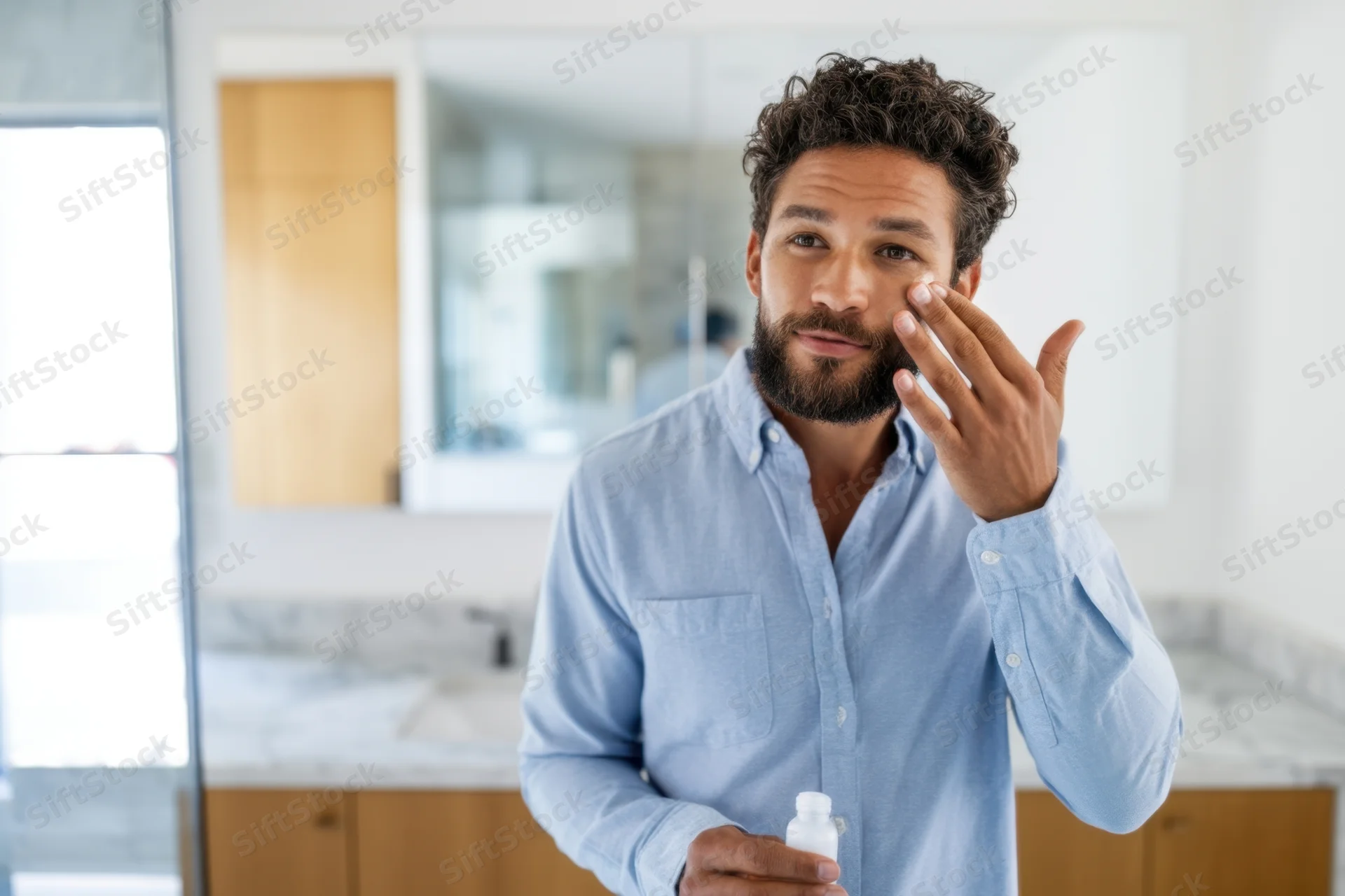 Young man applying moisturizer to his face in a modern bathroom