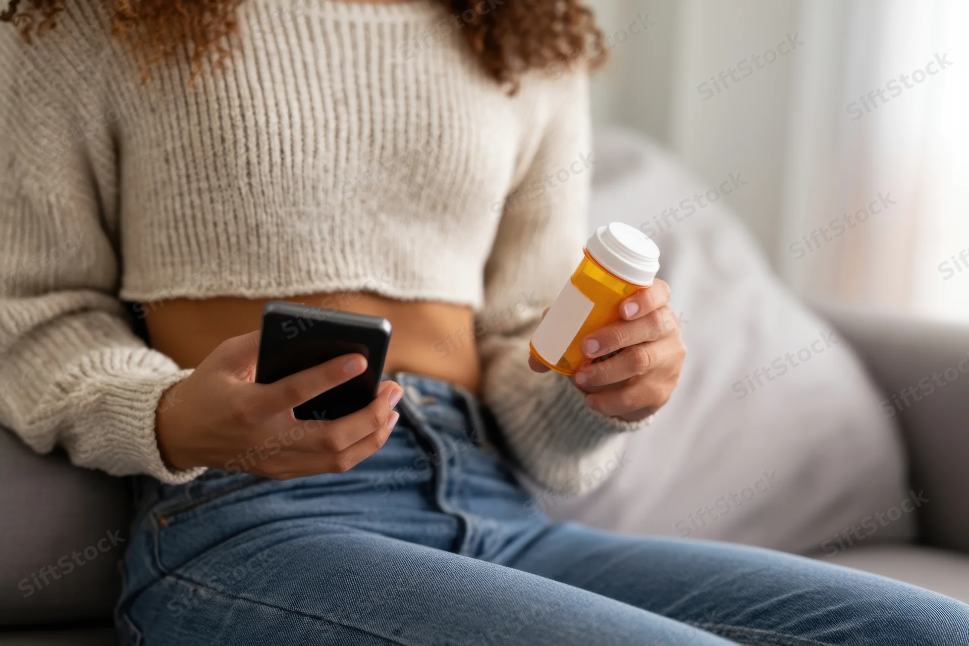 Young woman sitting on couch holding smartphone and prescription bottle