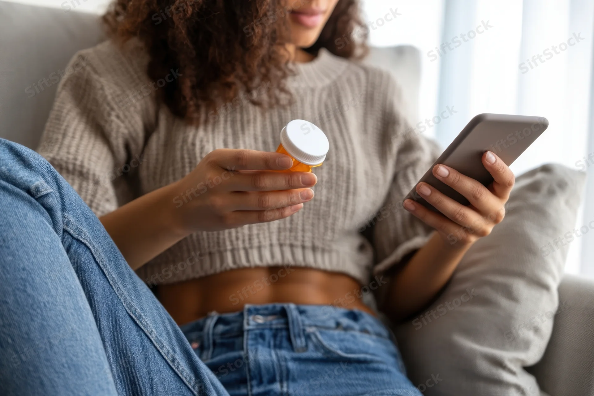 Young woman sitting on sofa holding medication bottle and smartphone
