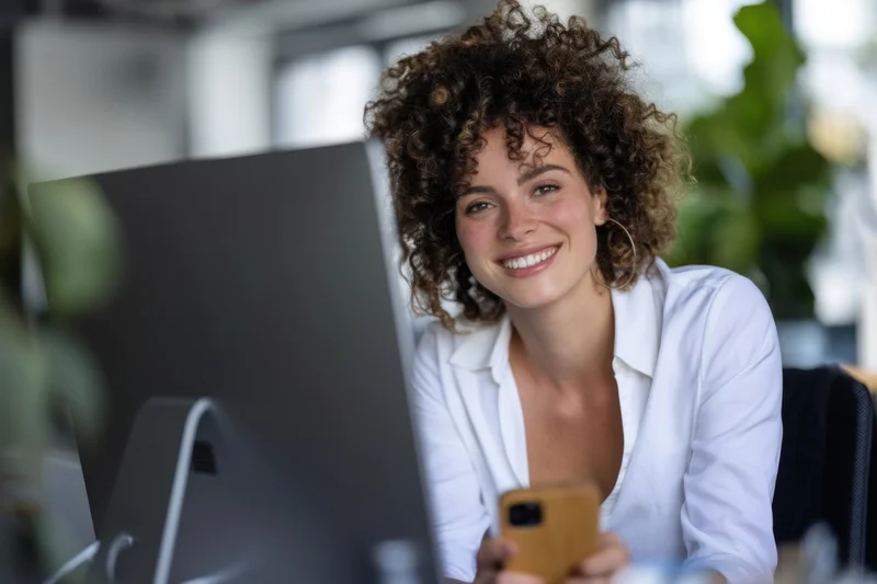 Young woman smiling at camera while using smartphone at office desk with computer monitor in front