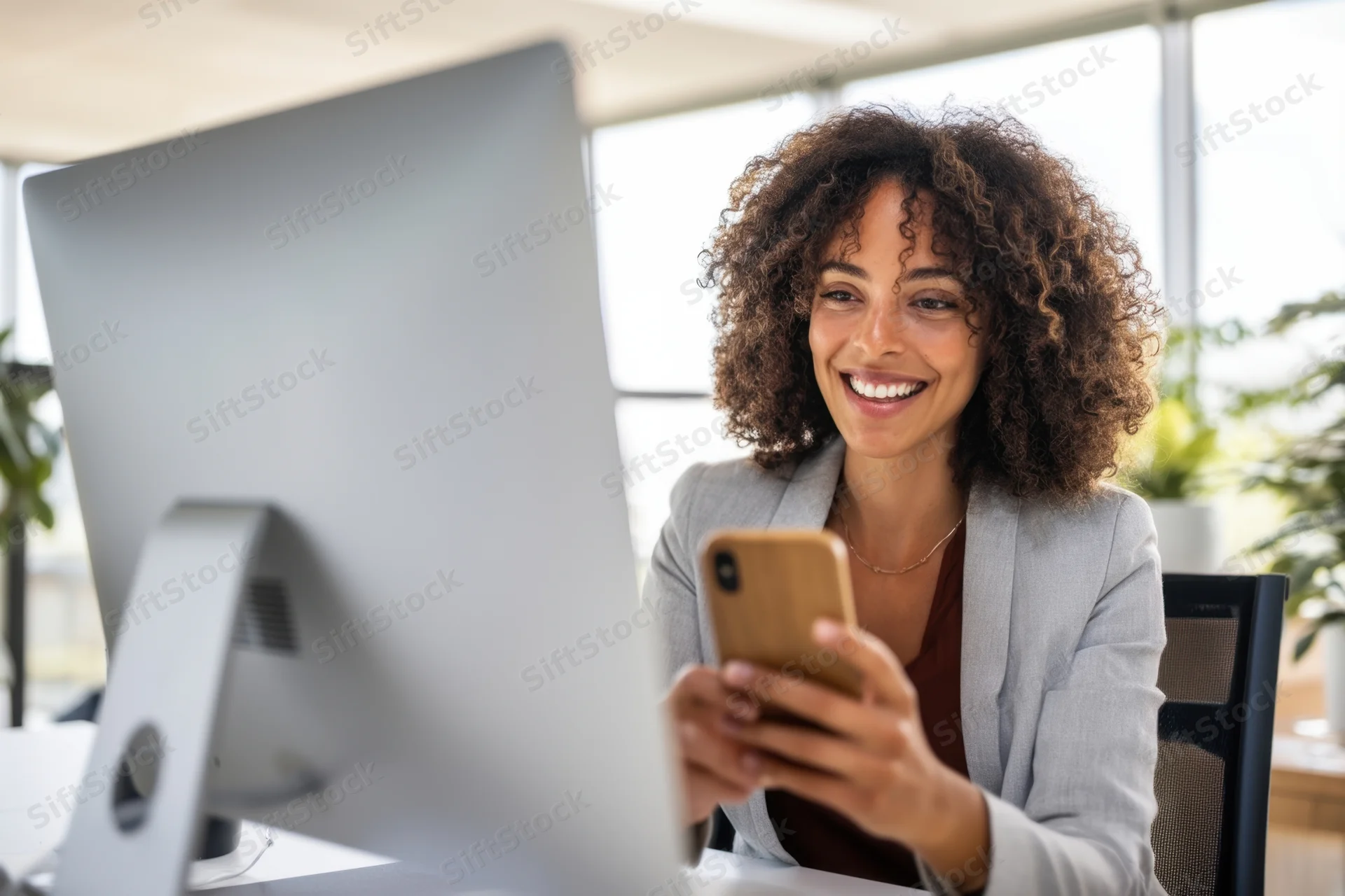 Young woman smiling while using smartphone at office desk with computer monitor