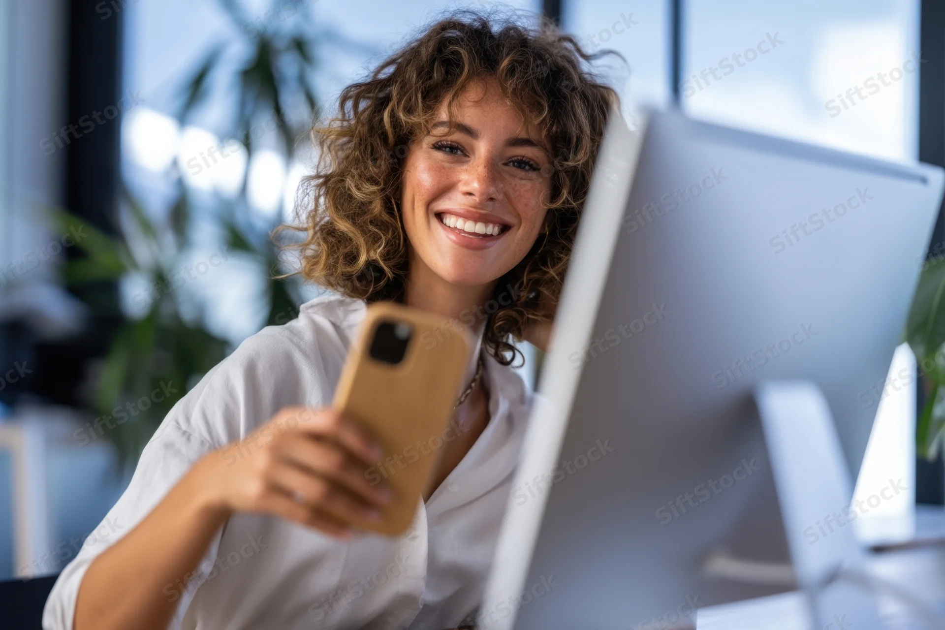 Young woman smiling while holding smartphone at office desk near computer monitor