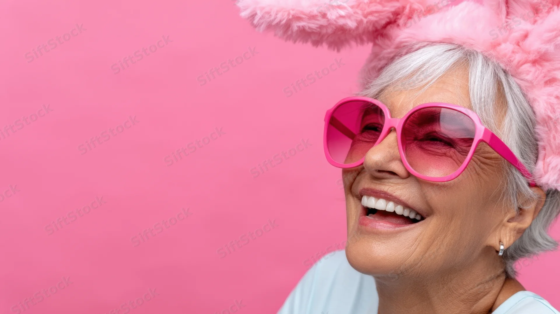 Smiling senior woman wearing pink bunny ears and sunglasses against pink background