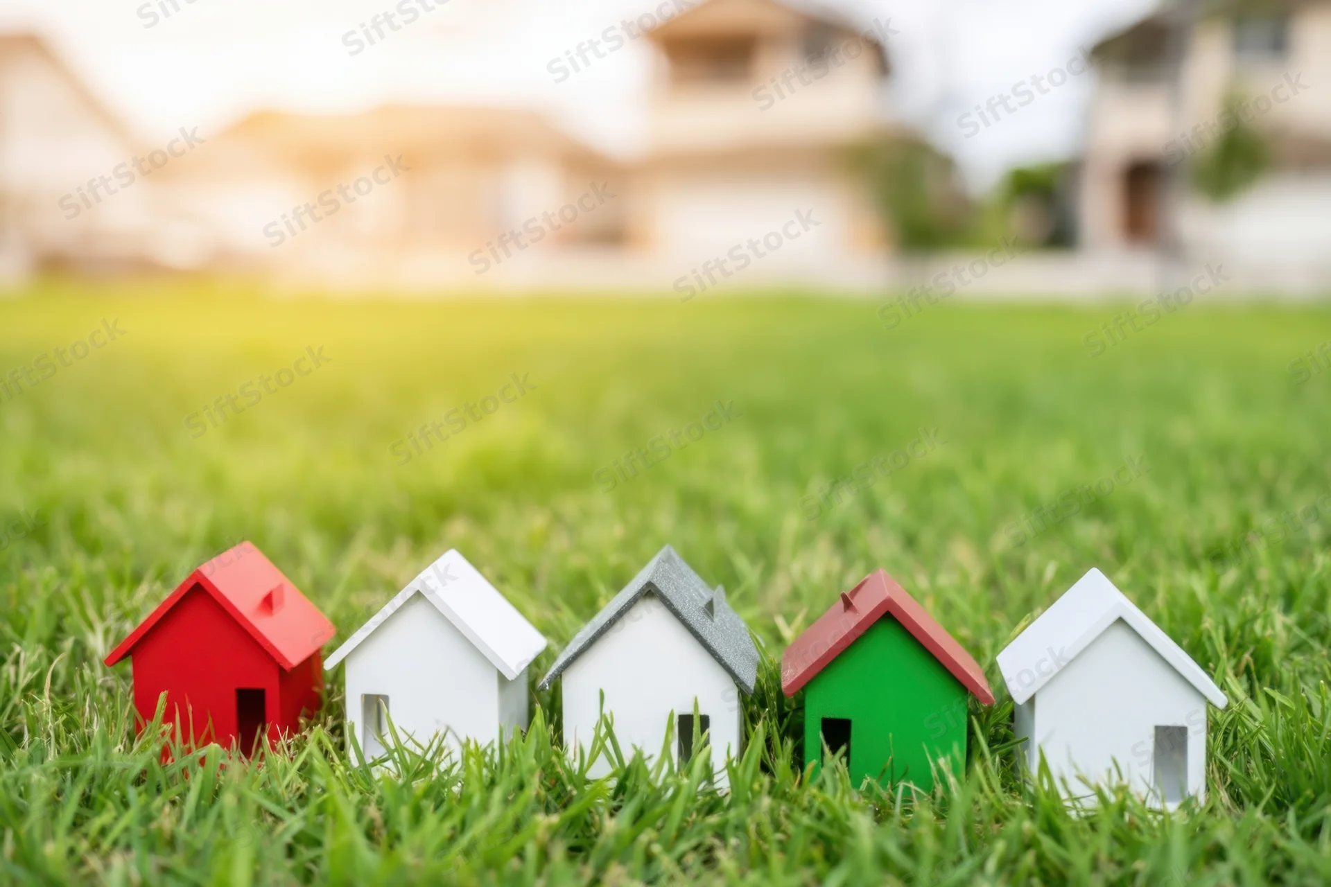 Miniature houses arranged in a row on a grassy lawn with blurred suburban homes in the background