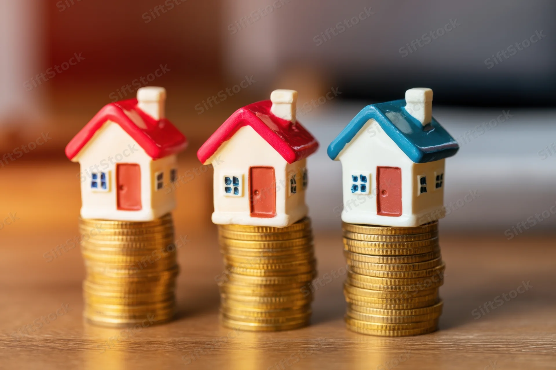 Three miniature houses on stacks of gold coins on a wooden surface